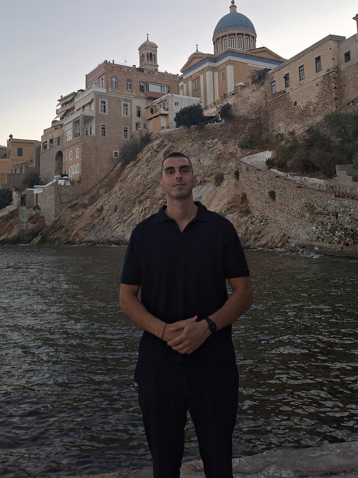 Christos Arseniou standing by the sea, with an island town and a church dome behind him